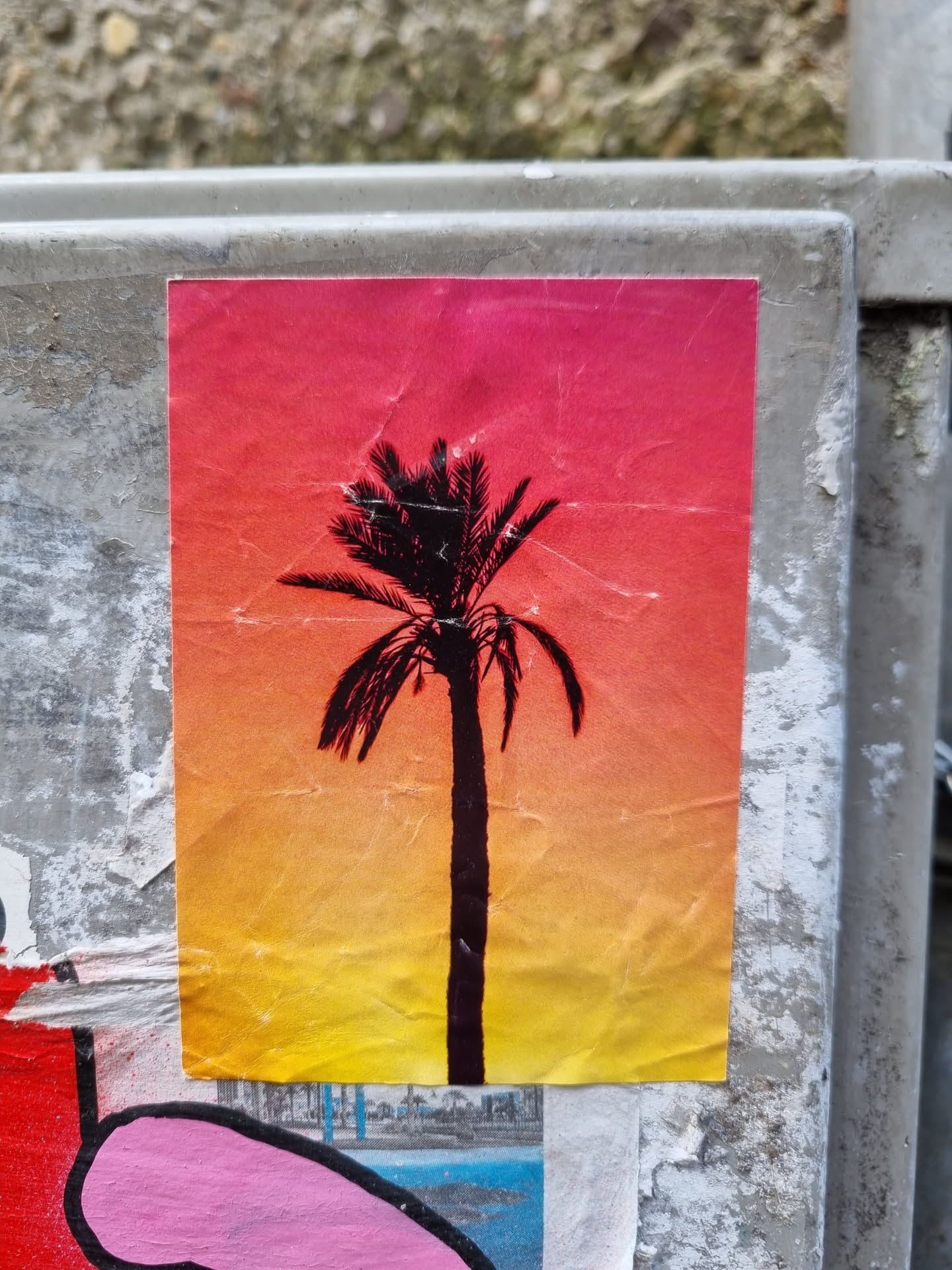 A sticker featuring a silhouette of a palm tree against a vibrant sunset backdrop. The colors transition from a deep pink/orange at the top to a warm yellow at the bottom. The sticker has a slightly creased and worn appearance, suggesting it may have been affixed outdoors for some time.
