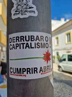 A sticker with a political message in Portuguese. It says "Derrube o capitalismo!" (Overthrow Capitalism!) and "Cumprir Abril" (Fulfill April), possibly referring to a political movement or event in April.  It includes a red stylized flower and a website address and Instagram handle.
