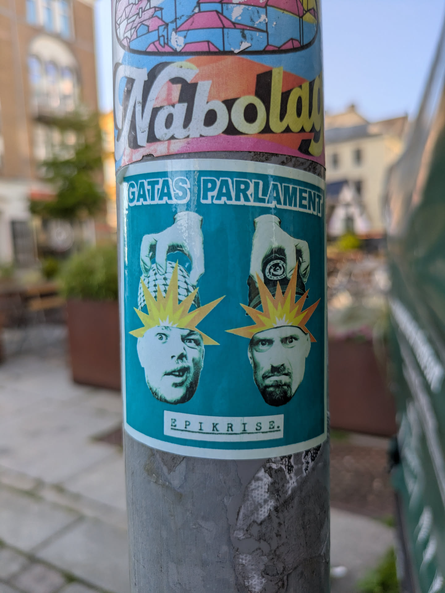 The sticker, predominantly teal blue, features two stylized male faces positioned at the bottom. Each face has a jagged, yellow-orange starburst graphic radiating from their head, suggesting an explosion of thought or ideas. Above the left face, a gloved hand holds a spiky, round object, possibly a grenade or a mind-bomb. The left face has an open mouth and wide eyes, appearing surprised or mid-expression. Above the right face, a hand holds a circular object that appears to be a seal or a symbol, with faint text 'SÄKRA FRAMTIDEN' visible on it. The right face has a beard and a more serious or contemplative expression. At the top of the sticker, the text 'GATAS PARLAMENT' is written in bold, white letters, and at the bottom, 'EPIKRISE.' is written in a more subtle, typewritten font.