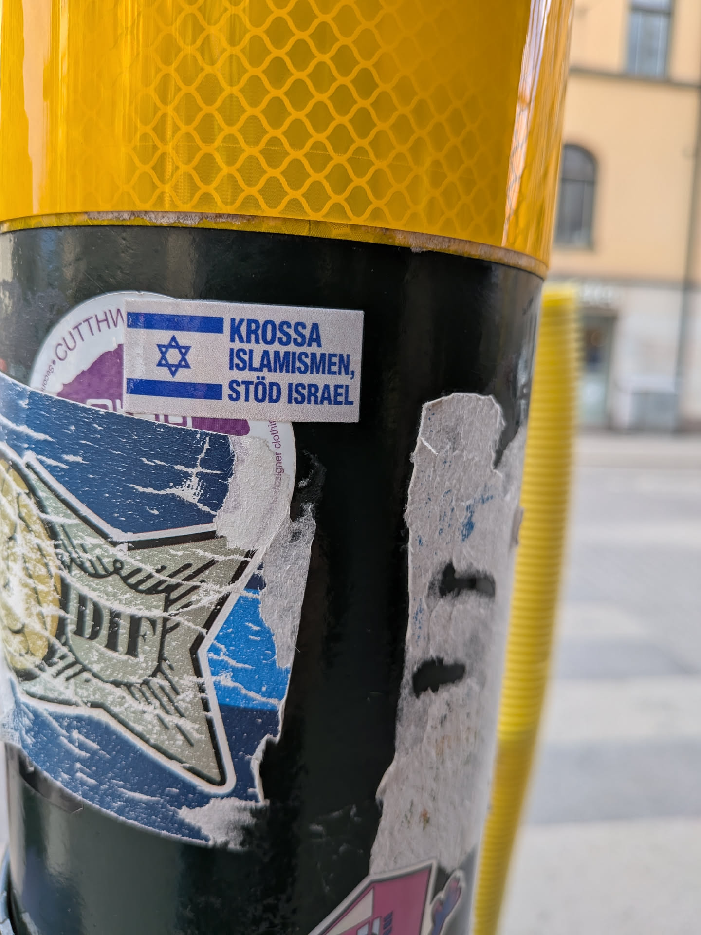 The sticker is rectangular, primarily white with blue text and imagery. On the left, it displays the flag of Israel, featuring two horizontal blue stripes at the top and bottom, with a blue Star of David centered on a white background. To the right of the flag, the text 'KROSSA ISLAMISMEN, STÖD ISRAEL' is printed in blue, sans-serif capital letters. The sticker shows signs of wear, with slightly peeling edges, and is affixed to a dark pole, partially overlapping other older, torn stickers. Above the pole is a yellow reflective surface.