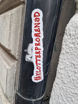 A red and white sticker on a black pipe with the text #Klotterpromenad. The sticker is rectangular with slightly rounded corners. The text is written in a stylized font.