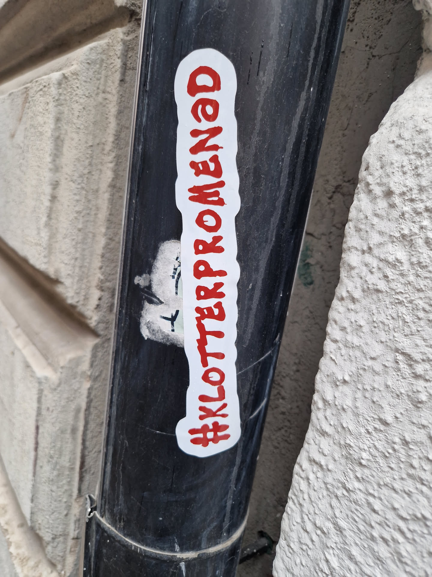 A red and white sticker on a black pipe with the text #Klotterpromenad. The sticker is rectangular with slightly rounded corners. The text is written in a stylized font.