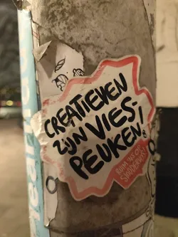 A sticker with Dutch text on it. The text says: "Creatieven zijn vies peuken" which translates to "Creatives are disgusting butts". There is also smaller text that says: "Ruim 'ns op stommevos!" which translates to "Clean up, you idiot!". The sticker is star-shaped and has a pink border. It is stuck on a gray pole.