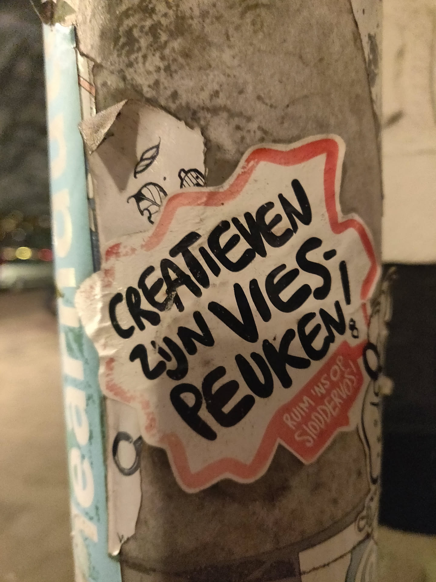 A sticker with Dutch text on it. The text says: "Creatieven zijn vies peuken" which translates to "Creatives are disgusting butts". There is also smaller text that says: "Ruim 'ns op stommevos!" which translates to "Clean up, you idiot!". The sticker is star-shaped and has a pink border. It is stuck on a gray pole.