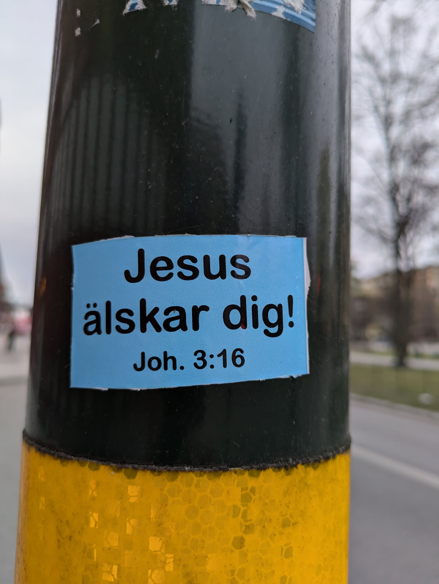 A blue rectangular sticker on a dark pole. The sticker contains the message "Jesus älskar dig!" (Jesus loves you!) followed by "Joh. 3:16" which is a reference to the Bible verse John 3:16.