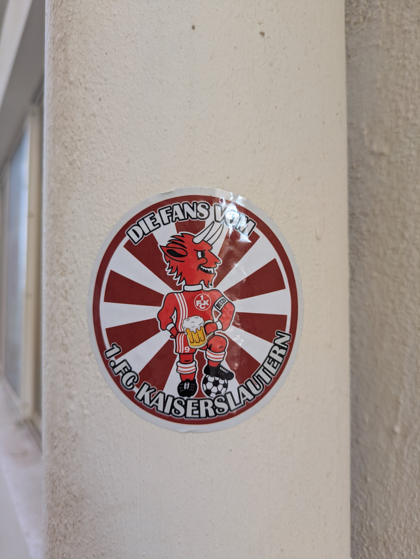 The sticker features a red devil mascot, a common symbol for the 1. FC Kaiserslautern football club. The devil is wearing the team's jersey with the number 9, holding a stein of beer in one hand and a soccer ball under its foot. The sticker is circular with red and white stripes in the background and text indicating it's a fan representation. "DIE FANS VOM 1.FC KAISERSLAUTERN".