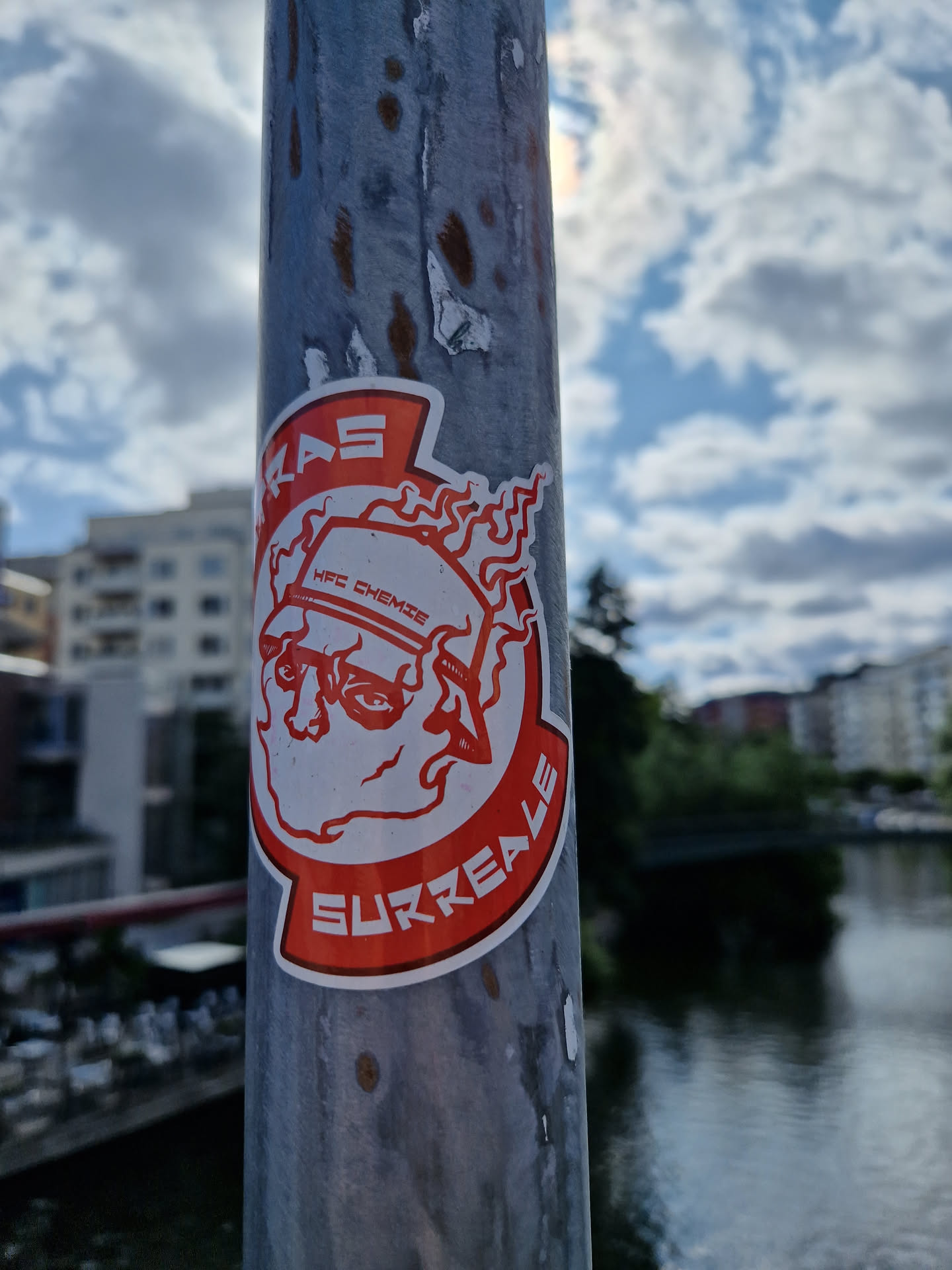 Red and white sticker featuring a surreal illustration of a face with flames, and the word 'Surreale' at the bottom. The text 'HPC CHEMISE' is visible on the top of the illustration. The sticker is attached to a gray metal pole outdoors, with a body of water visible in the background.