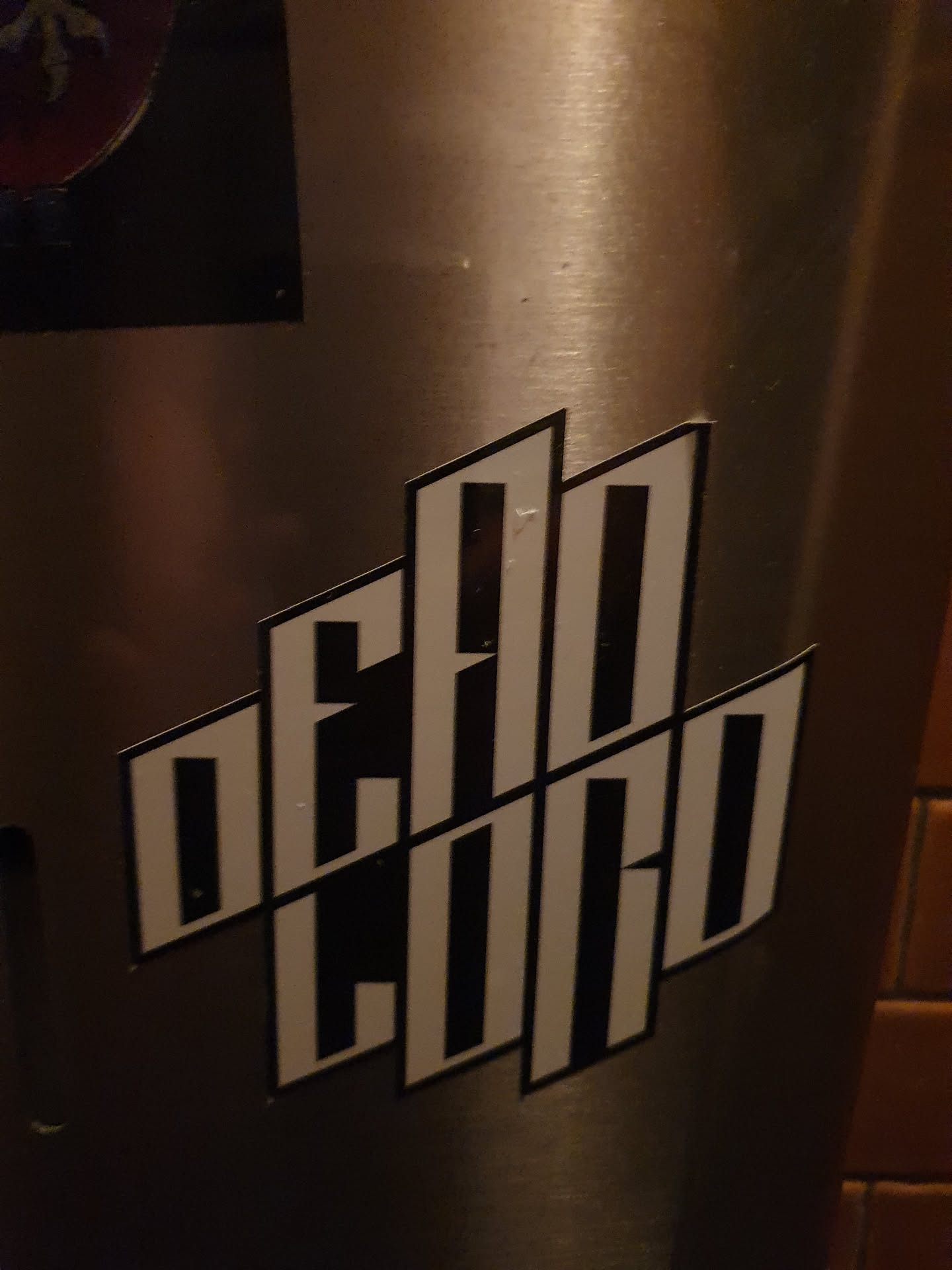 A stylish black and white sticker with the text "Dead Club" arranged in a geometric pattern. The sticker is affixed to a metallic surface.