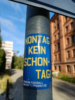 A blue sticker on a pole says that Monday is not a beautiful day, but a football day. The website www.lsv-tempo90ev.de is also mentioned in smaller text. The sticker is a great example of street art and German culture. It is a simple design but very effective in its message.