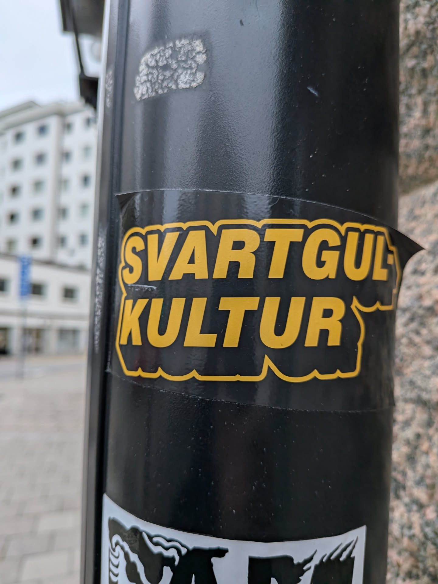 The sticker is predominantly black with bold, yellow lettering. The text is arranged in two lines: 'SVARTGUL' on the top line and 'KULTUR' on the bottom line. The letters have a thick, black outline, and the yellow fill has a slightly textured appearance. The sticker has a wavy, irregular edge, suggesting it's a die-cut sticker. It appears to be adhered to a black cylindrical pole.