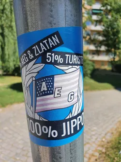 A blue sticker with a stylized American flag featuring the letters AEG.  The text on the sticker reads "AEG & Zlatan 51% Turister" at the top and "100% JIPPO" at the bottom. The sticker is affixed to a metal pole.