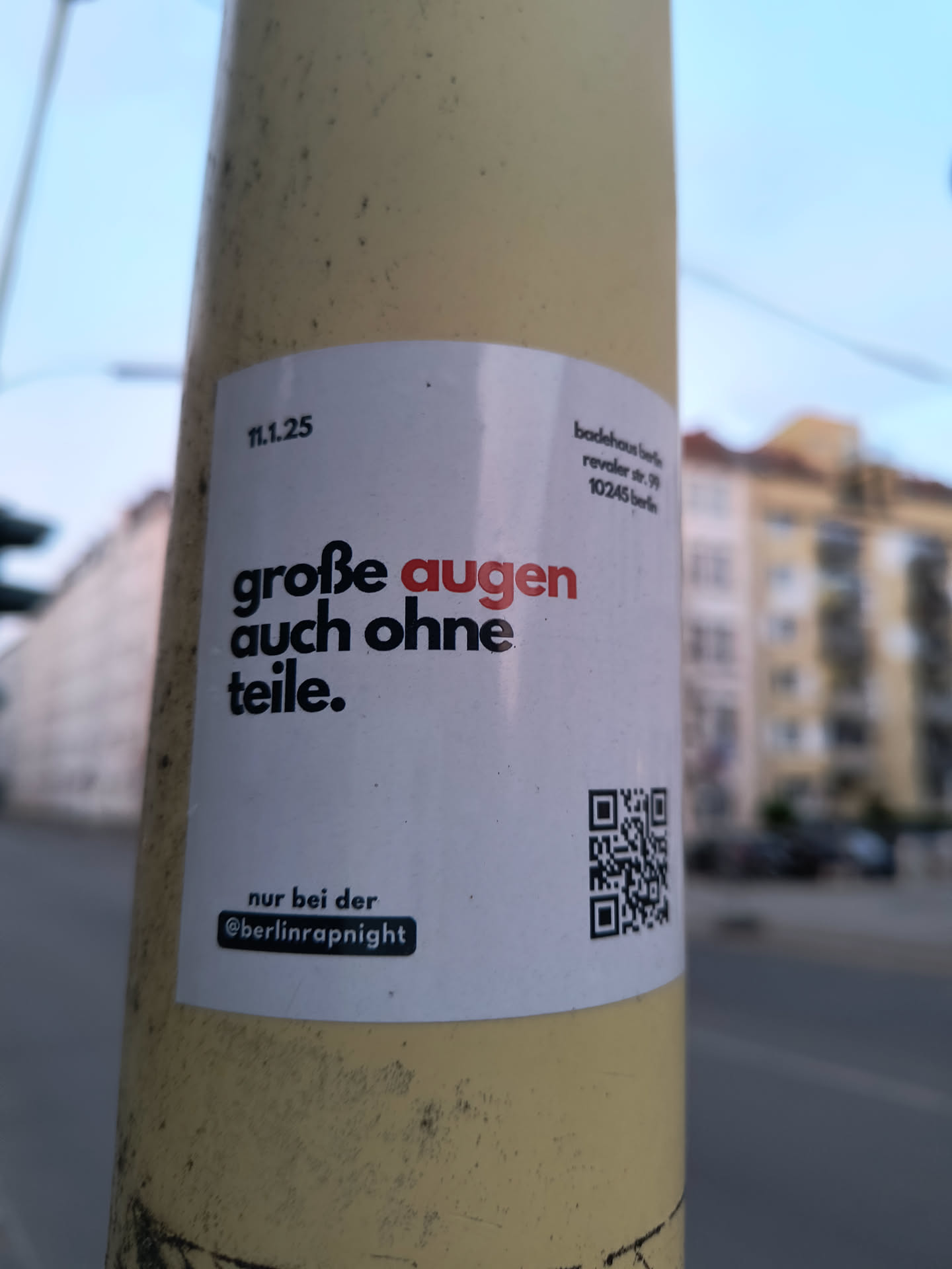 A white rectangular sticker with black and red text, adhered to a pole. The sticker reads "große augen auch ohne teile." with "augen" highlighted in red. Below this, it states "nur bei der @berlinrapnight". A QR code is located on the right side of the sticker. The sticker promotes Berlin Rap Night and includes their Instagram handle.