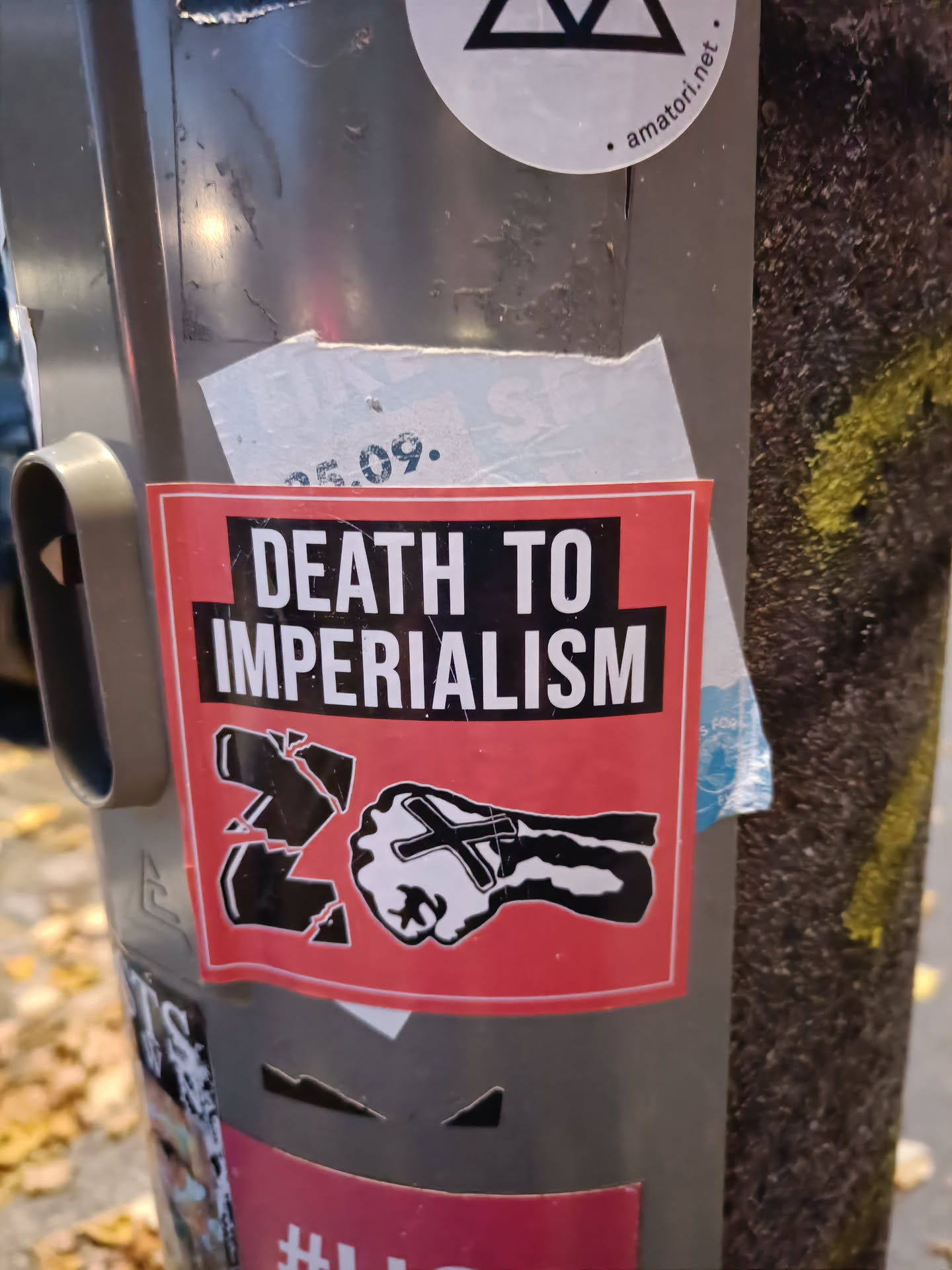 A red sticker with a black fist and the words "Death to Imperialism". The fist is broken into pieces. The sticker is on a grey pole.