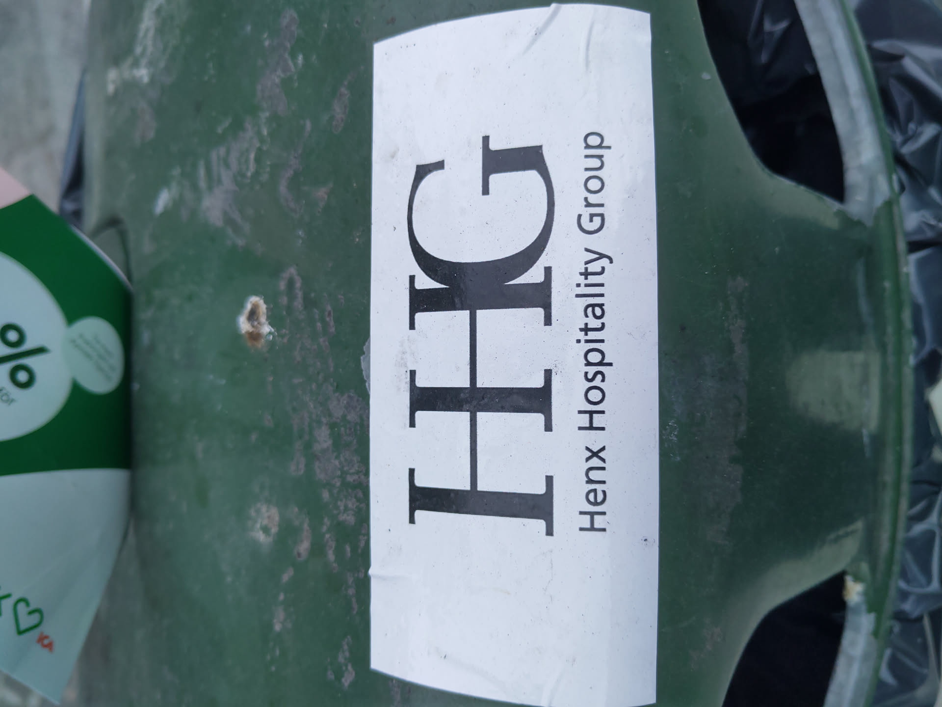 A white sticker with the letters HHG and the text Henx Hospitality Group is affixed to a dark green surface. The sticker shows some signs of wear.
