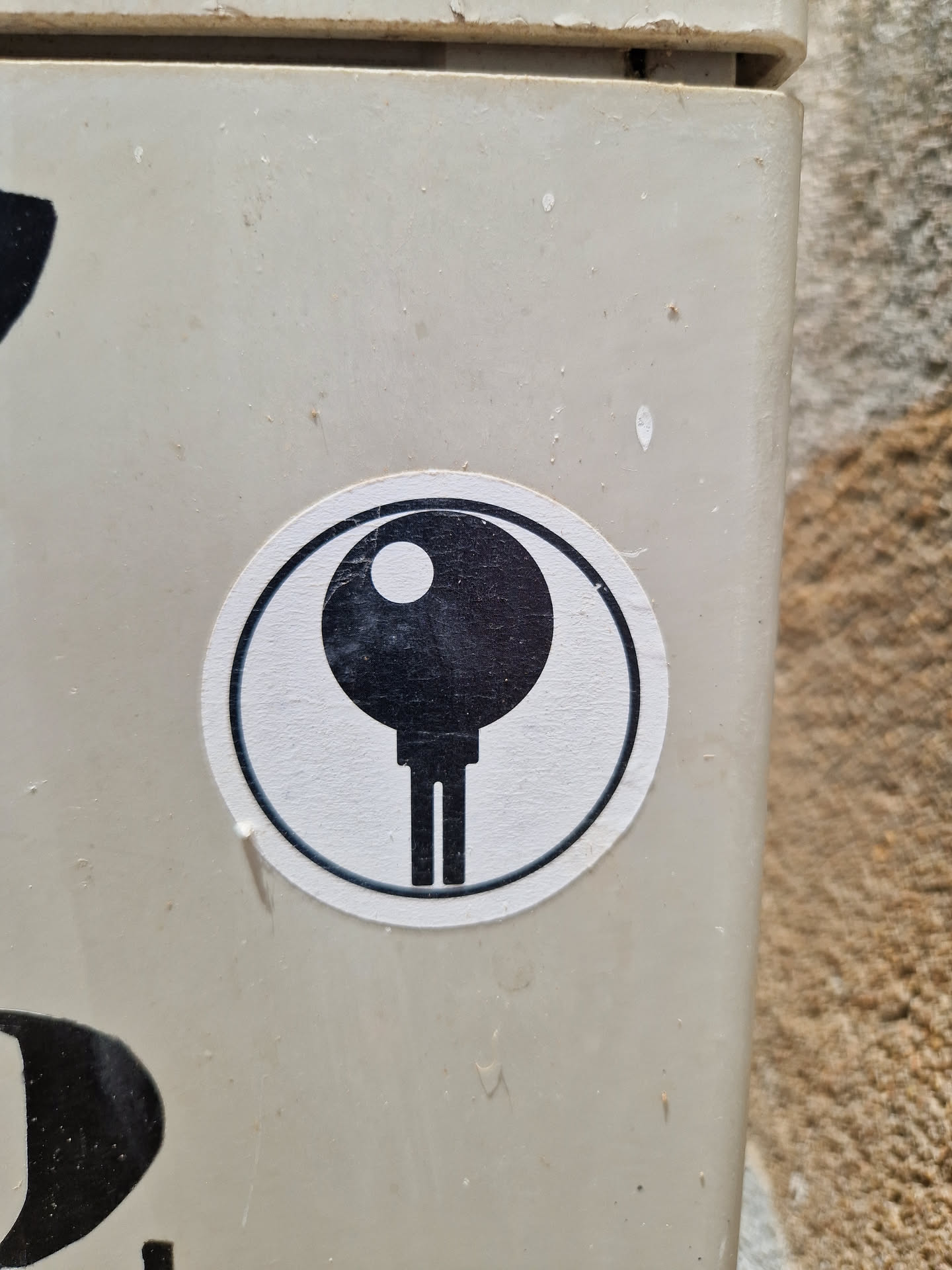 A round sticker with a black and white minimalist design. The symbol in the center resembles a stylized key or a simple machine part.  It's affixed to a light beige surface.