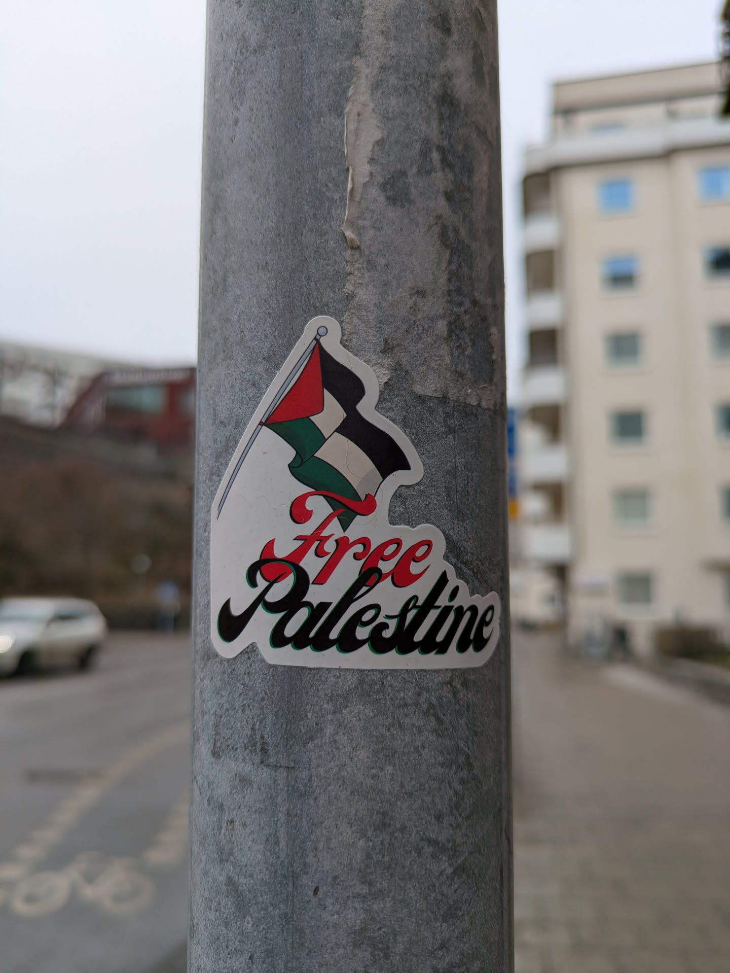 A sticker featuring the Palestinian flag with the words "Free Palestine" is affixed to a gray metal pole. The flag is illustrated in a cartoon style, with the black, white, and green sections waving from a red pole. The words "Free Palestine" are written in a decorative script below the flag, with "Free" in red and "Palestine" in black.