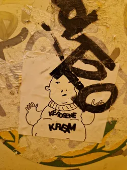A sticker featuring a cartoon drawing of a chubby character wearing a sweater with the words "Kerosene Kream" written on it. The drawing is in black ink on white paper. The sticker is attached to a wall with other graffiti and markings.