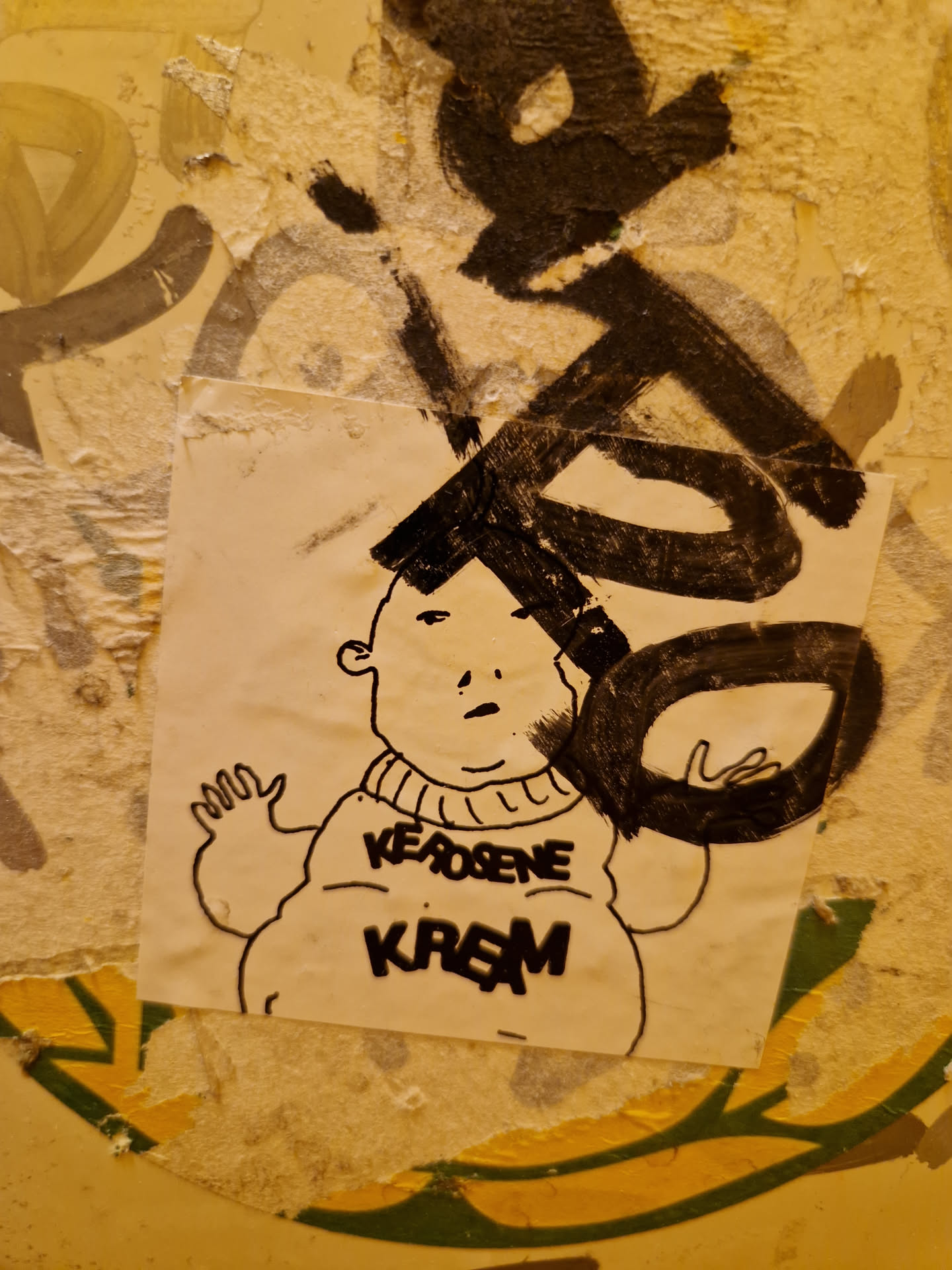 A sticker featuring a cartoon drawing of a chubby character wearing a sweater with the words "Kerosene Kream" written on it. The drawing is in black ink on white paper. The sticker is attached to a wall with other graffiti and markings.