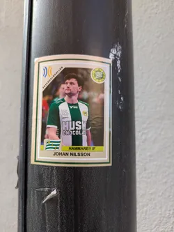 The sticker is a trading card style image of Johan Nilsson wearing his green and white Hammarby IF jersey with the Husk chocolate logo. The text "Johan Nilsson" and "Hammarby IF" is visible on the card. The background shows part of the Erikdalshallen arena.