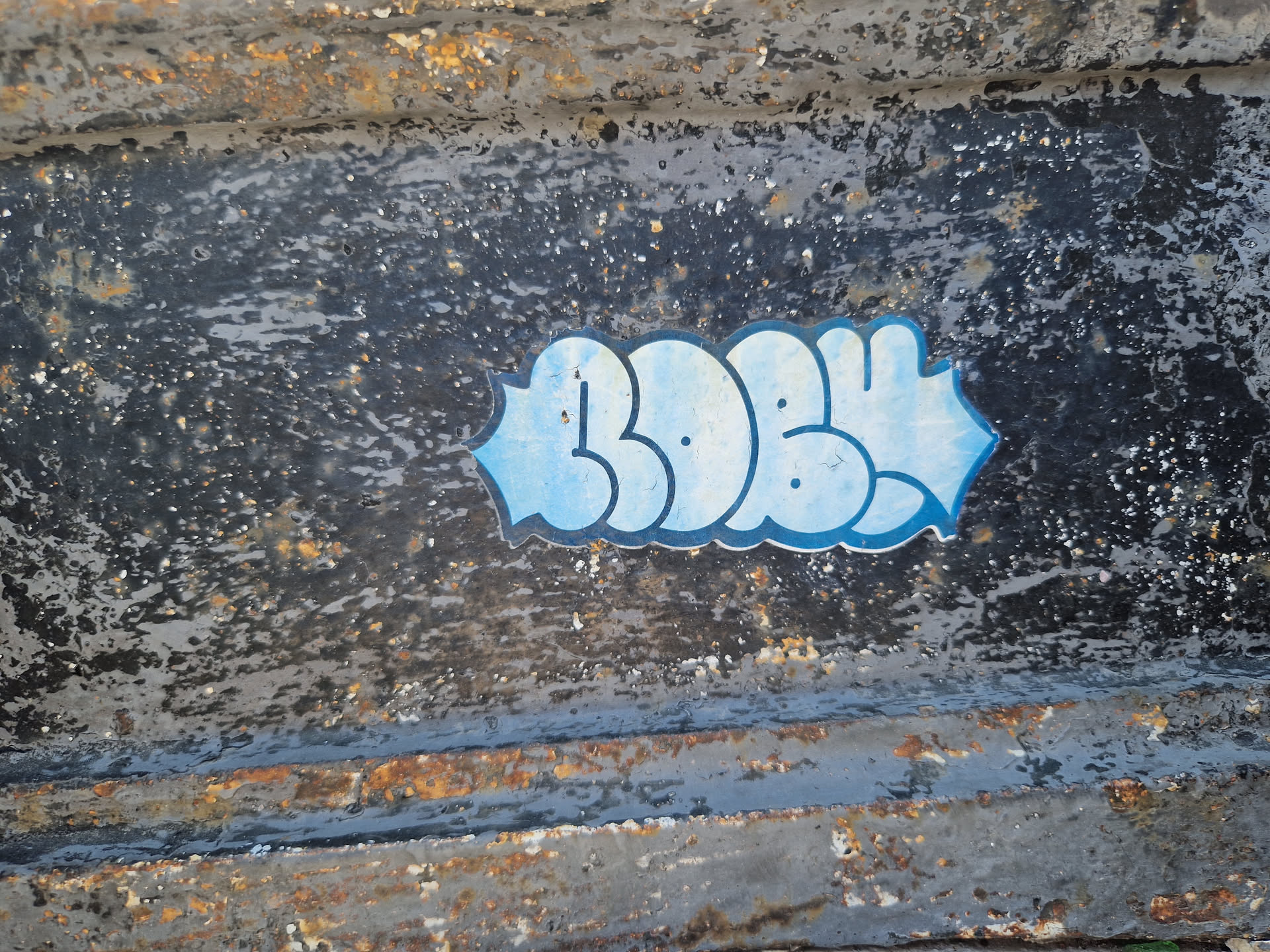 A light blue sticker featuring stylized bubble letters that spell out "BOBY" is affixed to a dark gray, heavily rusted metal surface. The sticker's edges are slightly irregular. The background is textured and shows significant signs of wear and rust.
