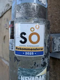 A rectangular sticker with rounded corners, featuring a clean, modern design. The background is white. At the top, a prominent logo displays a large black 'S' followed by a large orange 'Ö', with two smaller orange circles above the 'Ö'. Below the logo, the text 'SVENSKA ÖLFRÄMJANDET' is written in black, sans-serif font. A horizontal yellow band spans the width of the sticker, containing the word 'Rekommenderar' in black, bold, sans-serif text. Below this, a blue band features the year '2025' in white, bold text, flanked by small white dots.