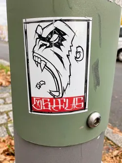 The sticker features a striking black and white illustration of an aggressive gorilla or ape head, viewed in profile, with its mouth wide open revealing sharp fangs and an angry expression. The art style is bold and graphic, characterized by thick black outlines and cross-hatching to denote shadow and texture, especially around the eyes and brow. The main body of the ape is white. Below the illustration, there is a prominent horizontal red banner with heavily stylized white text. The entire design is contained within a thin black border, and the sticker itself is rectangular with slightly rounded corners, showing minor wear and a slight lift at the top edge.