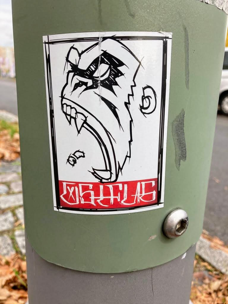 The sticker features a striking black and white illustration of an aggressive gorilla or ape head, viewed in profile, with its mouth wide open revealing sharp fangs and an angry expression. The art style is bold and graphic, characterized by thick black outlines and cross-hatching to denote shadow and texture, especially around the eyes and brow. The main body of the ape is white. Below the illustration, there is a prominent horizontal red banner with heavily stylized white text. The entire design is contained within a thin black border, and the sticker itself is rectangular with slightly rounded corners, showing minor wear and a slight lift at the top edge.