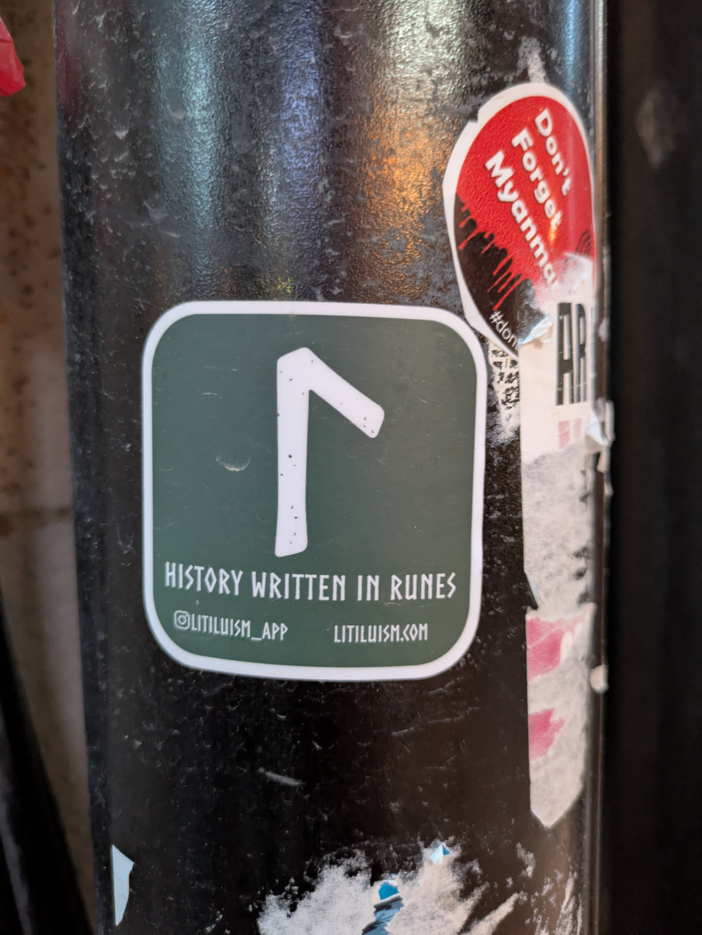 The central sticker is square with rounded corners and a thick white border. The background is a deep forest green. In the center, a white rune symbol resembling an upside-down 'F' or a stylized 'R' is prominently displayed. Below the rune, in white, sans-serif capital letters, is the text 'HISTORY WRITTEN IN RUNES'. Beneath this, in smaller white text, are two items: an Instagram handle '@LITILUISM_APP' and a website 'LITILUISM.COM'. The sticker appears to be affixed to a dark, textured cylindrical surface, possibly a pole, which shows signs of wear and other stickers. To the upper right, a red and black oval sticker with the text 'Don't Forget Myanmar' is partially visible.