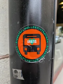 A round sticker attached to a black pole features an orange bus icon with the word 'GRATIS' (meaning 'free' in Swedish) in green within the bus's body. The text 'ÅTERSTÄLL KOLLEKTIVTRAFIKEN' encircles the bus, which translates to 'Restore Public Transport'. The sticker is slightly worn.