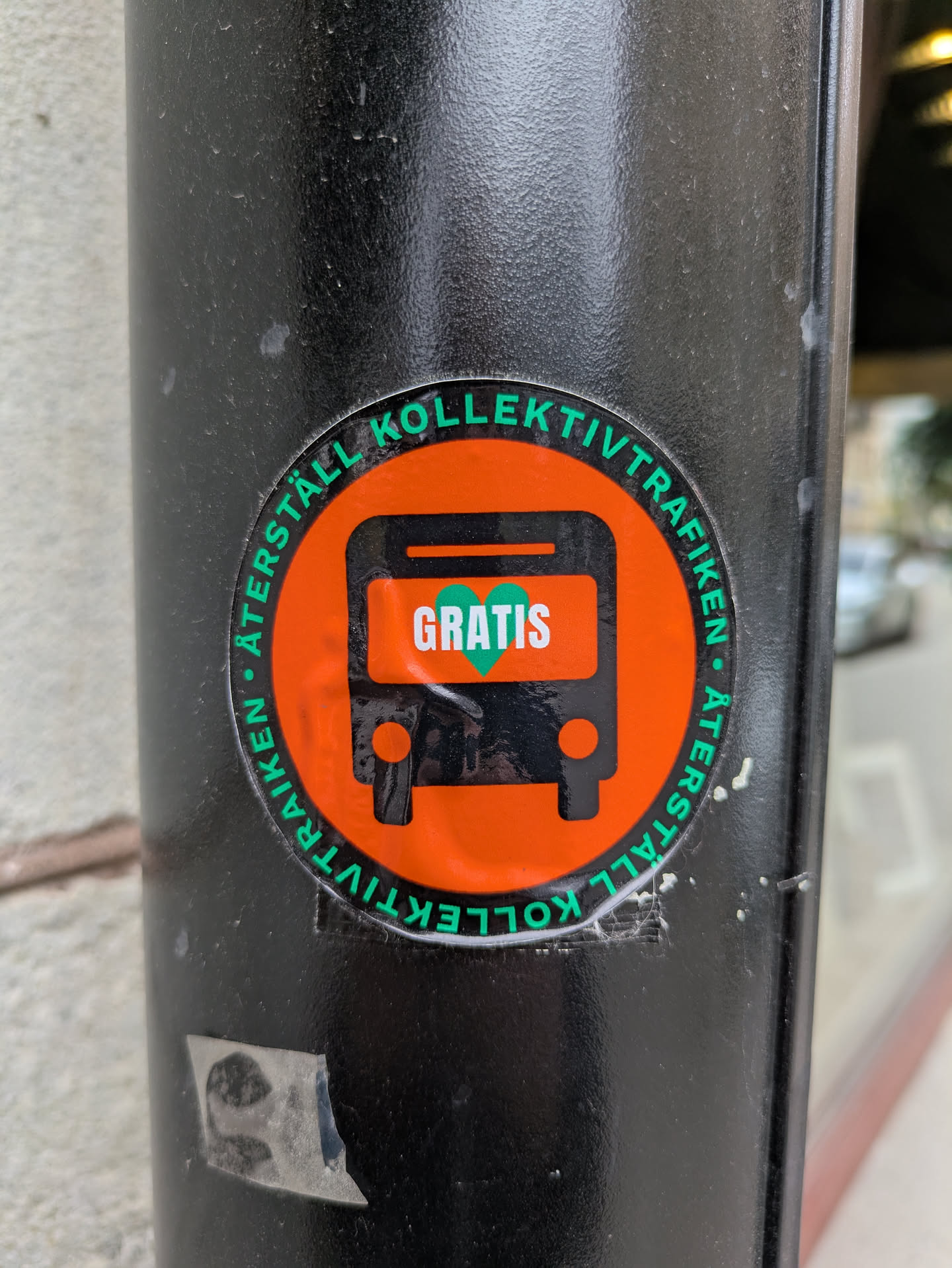 A round sticker attached to a black pole features an orange bus icon with the word 'GRATIS' (meaning 'free' in Swedish) in green within the bus's body. The text 'ÅTERSTÄLL KOLLEKTIVTRAFIKEN' encircles the bus, which translates to 'Restore Public Transport'. The sticker is slightly worn.