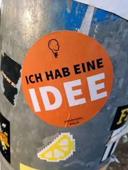 An orange round sticker with the text "ICH HABE EINE IDEE" in black and white, meaning "I have an idea" in German.  There is a small logo of a head with a lightbulb above it. The text "erfinderladen BERLIN" is printed at the bottom in a smaller font size.