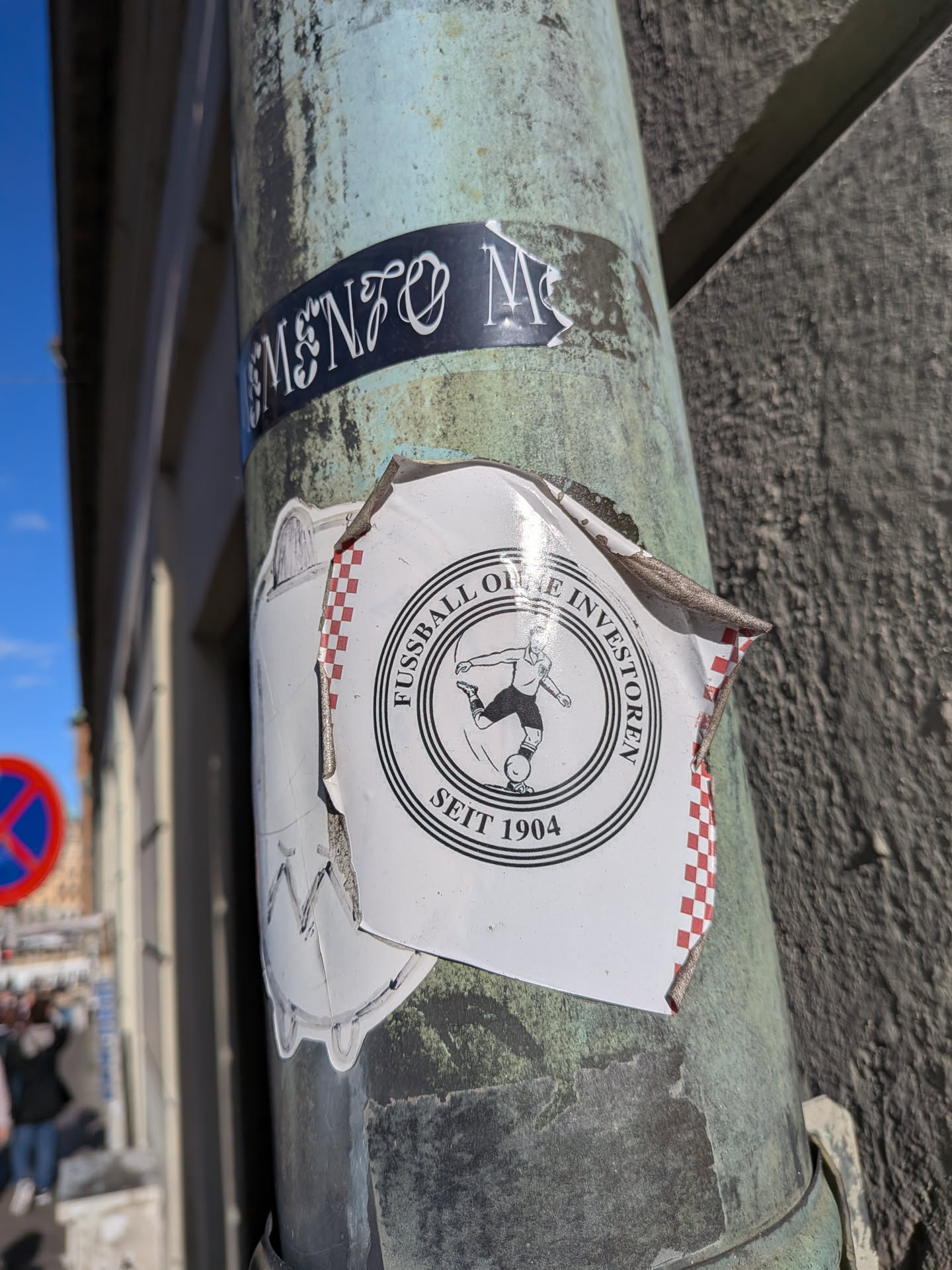 A peeling white sticker posted on a weathered pole, layered over another drawing. It displays a black circular crest featuring a vintage illustration of a footballer kicking a ball. Surrounding the central figure within concentric circles are the German words 'FUSSBALL OHNE INVESTOREN' along the top curve and 'SEIT 1904' at the bottom. The vertical borders of the sticker have a red-and-white checkered pattern.