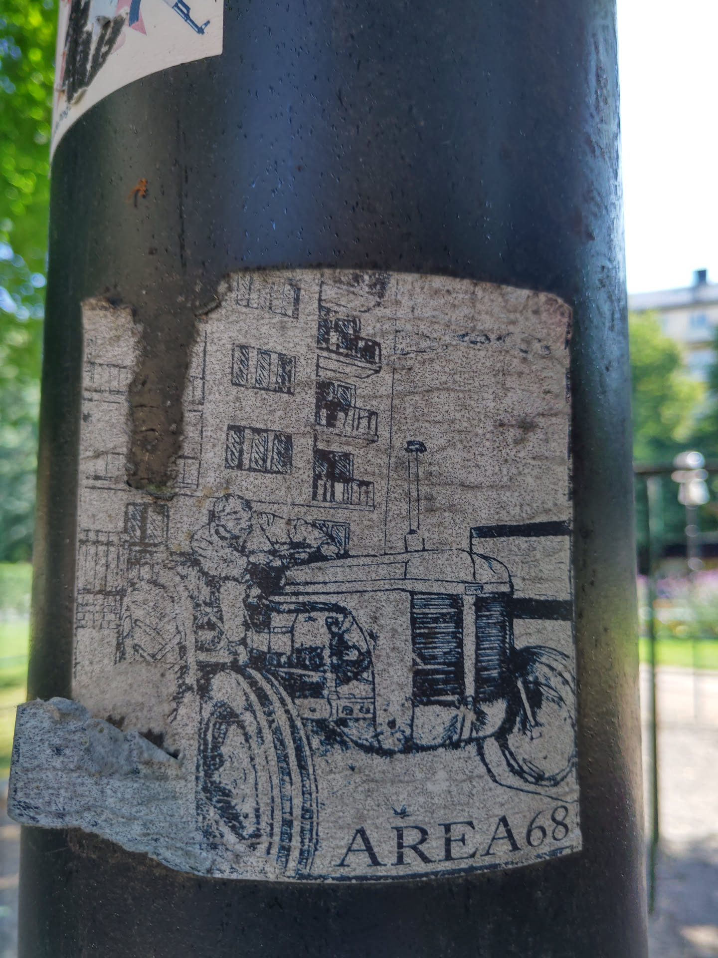 Black and white illustration of a tractor in front of a building. The sticker is on a dark pole. The text AREA68 is visible at the bottom.