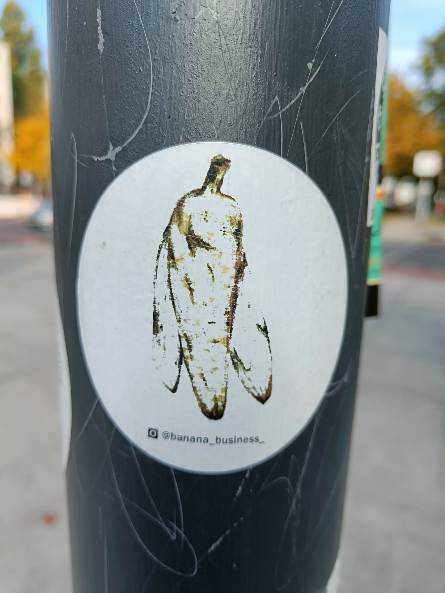 A round sticker featuring a stylized illustration of a banana. The banana is depicted in muted tones of yellow and brown, giving it an aged or vintage appearance. The style is simple and somewhat abstract, with a focus on shape and form rather than photorealistic detail.  The sticker has the Instagram handle @banana_business_ printed at the bottom.