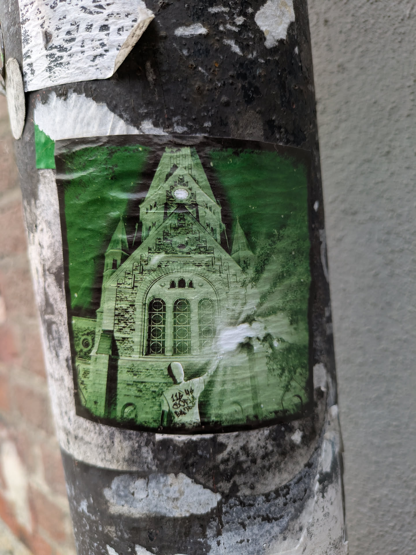This sticker features a monochrome image of a person seemingly radiating light in front of a church. The image is in shades of green and has a slightly faded quality, adding to its mysterious atmosphere. The text on the figure's back is partially visible but illegible. The sticker shows signs of wear, indicating it has been exposed to the elements for some time.