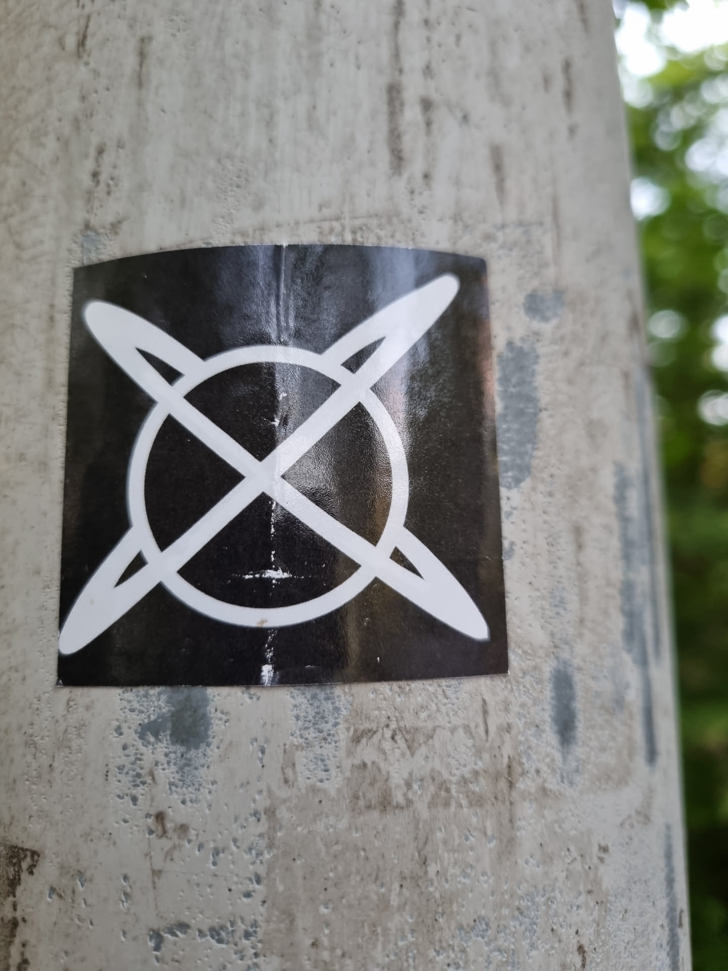 A square black and white sticker with a symbol that looks like a circle with lines crossing through it. The sticker is attached to a grey pole.