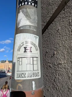 Street sticker A light-colored rectangular sticker featuring black line art. At the top, the words 'HOLY BURGER' curve above a stylized, bold letter 'H'. Beneath is a detailed line illustration depicting the storefront of the burger restaurant, complete with an awning, window frames, and entrance. At the bottom, the words 'DEZ ANOS' ('Ten Years' in Portuguese) are printed in an outlined, sans-serif font.