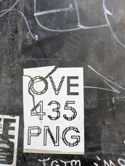 A cracked sticker adhered to a dark surface reads 'OVE 435 PNG'. The sticker has a pixelated design, giving it a retro, digital look. It appears to be slightly damaged, with a crack running through the left side of the sticker.
