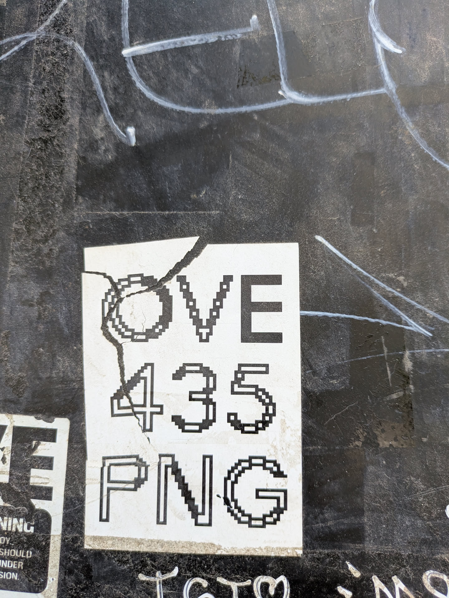 A cracked sticker adhered to a dark surface reads 'OVE 435 PNG'. The sticker has a pixelated design, giving it a retro, digital look. It appears to be slightly damaged, with a crack running through the left side of the sticker.