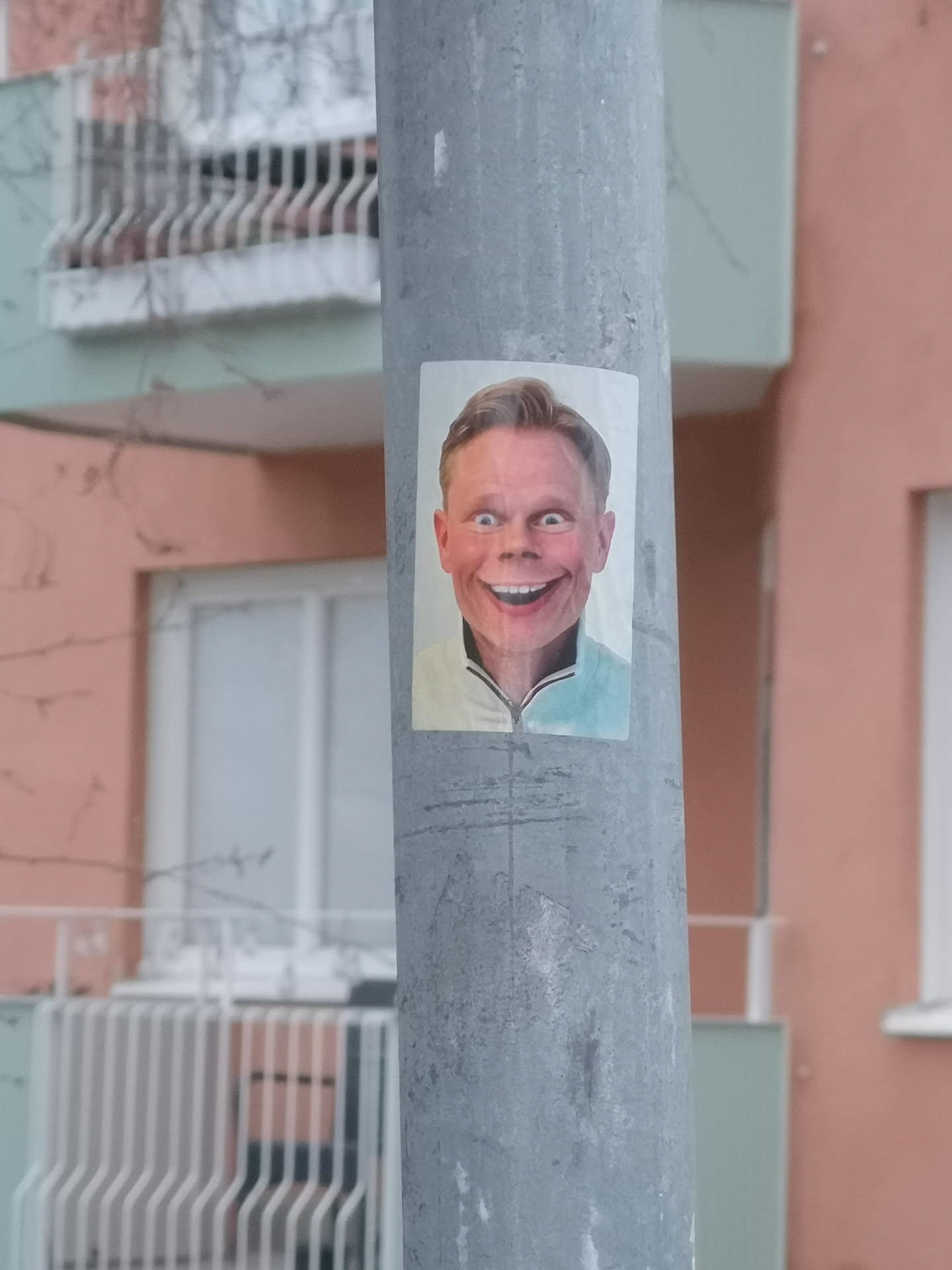 The sticker is a rectangular, vertically oriented image featuring a man's headshot. He has light brown hair, fair skin, and a very wide, almost exaggerated, toothy smile. His blue eyes are wide open, contributing to an intense or slightly unsettling expression. He is wearing a light-colored, possibly mint green or light blue, collared shirt with a zipper at the neck. The background of the portrait is plain white. The sticker is affixed to a grey, textured metal pole.
