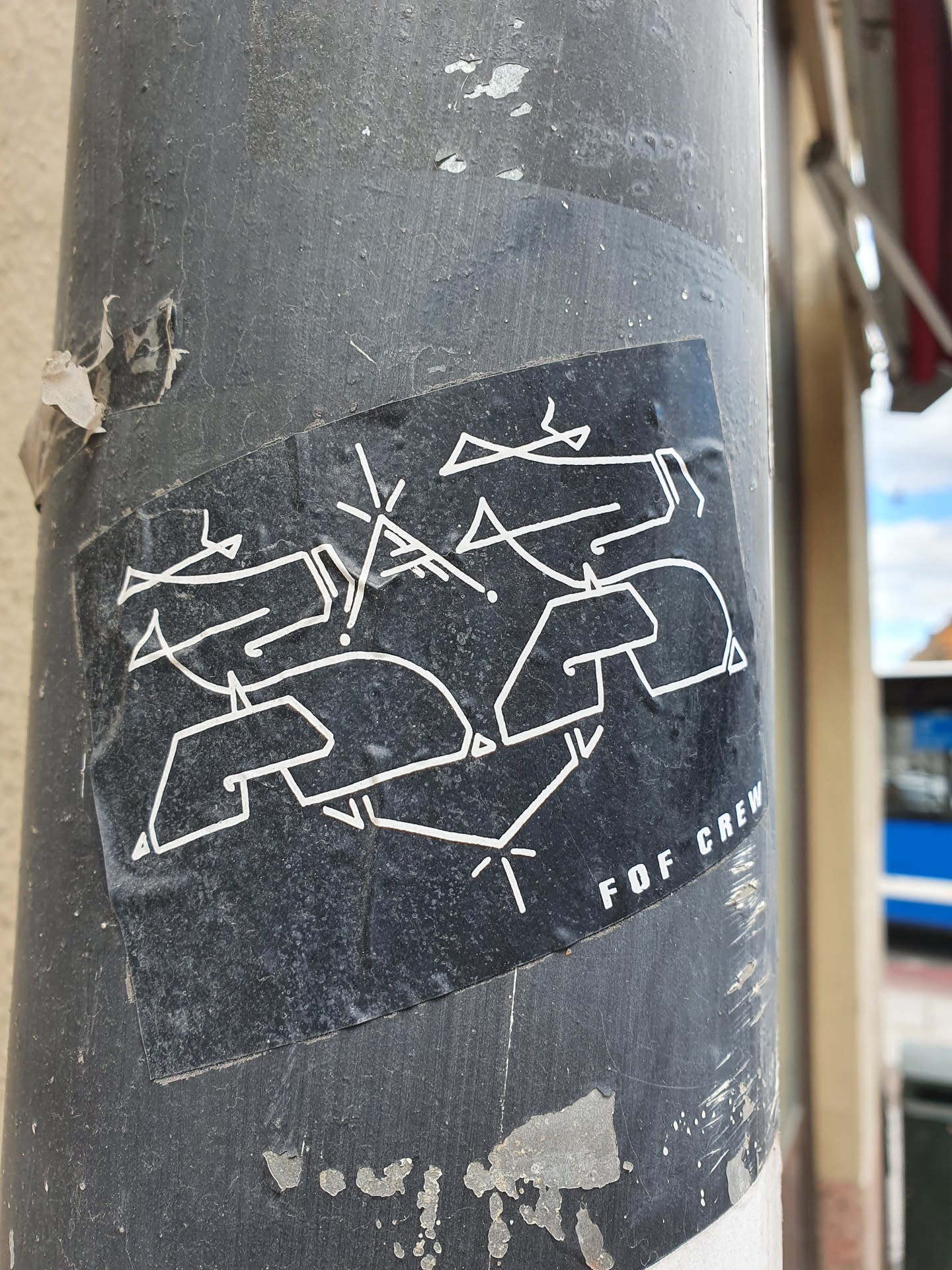 The sticker features a stylized graphic that appears to be a logo or tag, predominantly in white against a black background.  The text "FOF CREW" is visible at the bottom right. The sticker shows some wear and tear, indicating it may have been placed outdoors for a while. It's stuck on a weathered gray pole.