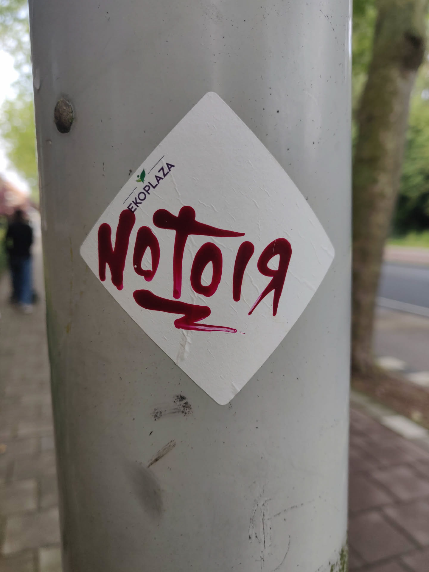A diamond-shaped sticker with the text \"NoTo19\" written in maroon paint. The sticker is affixed to a gray pole.  A small EKOPLAZA logo is visible in the upper part of the sticker.