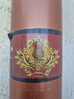 A sticker featuring an ornate design with a central hop cone, surrounded by decorative flourishes. The text at the bottom reads "Augusta Vindelicorum". The sticker is affixed to a brown cylindrical object.