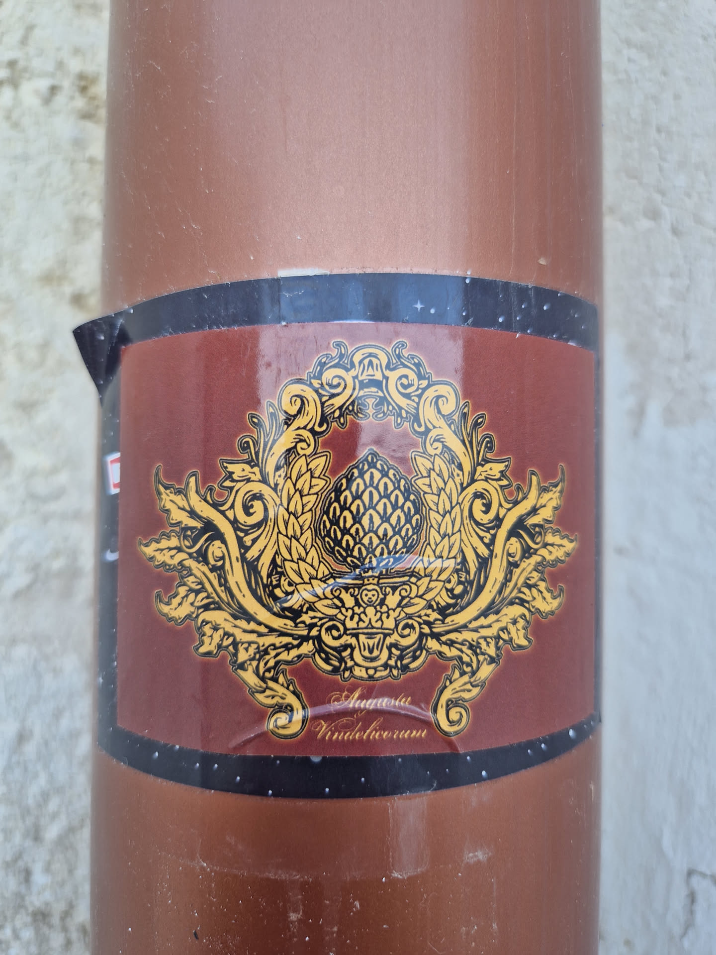 A sticker featuring an ornate design with a central hop cone, surrounded by decorative flourishes. The text at the bottom reads "Augusta Vindelicorum". The sticker is affixed to a brown cylindrical object.