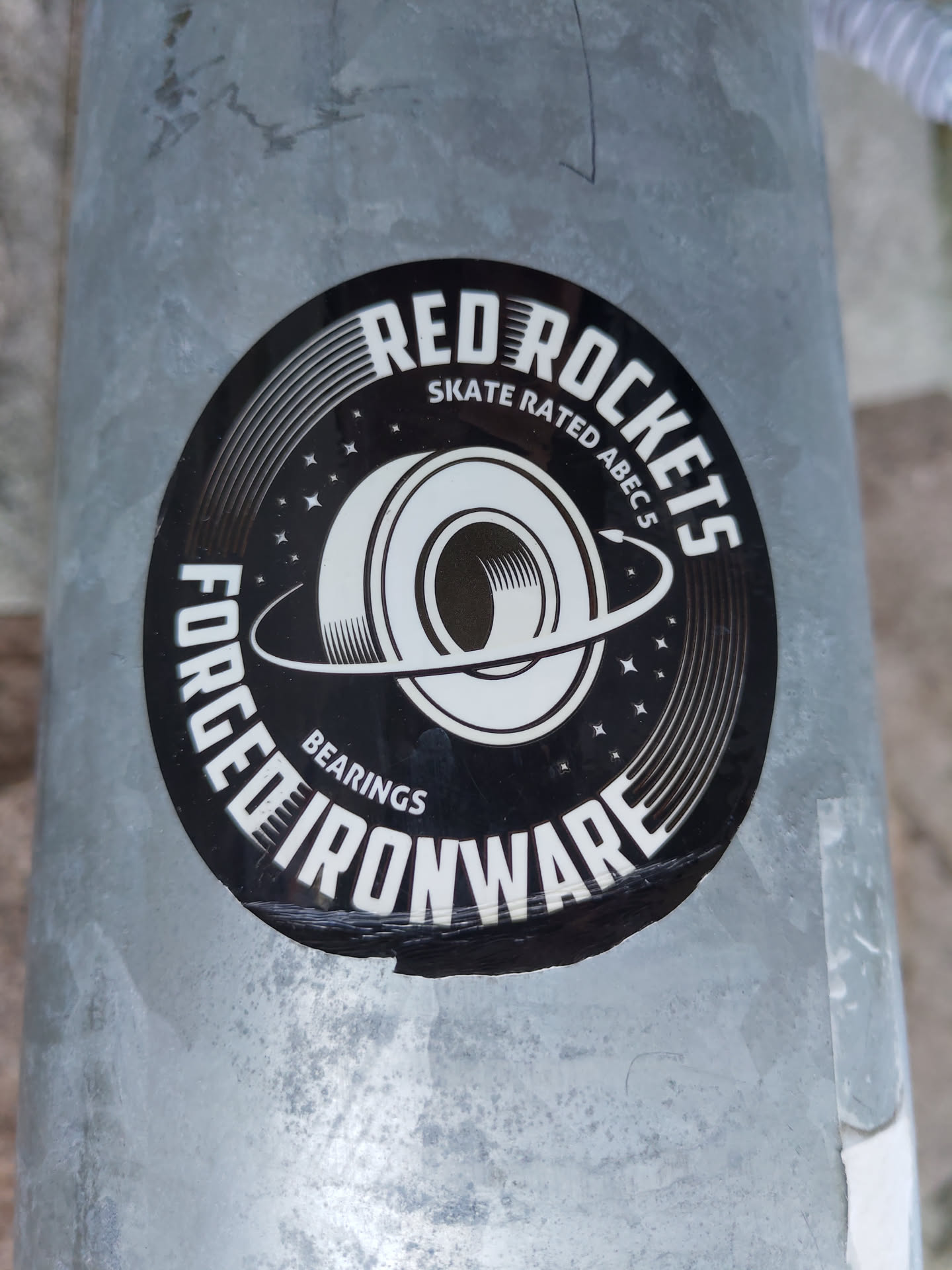 A black and white circular sticker with the text "Red Rockets Forged Ironware" written around a graphic of a bearing. The text "Skate Rated ABEC 5" is written in smaller font above the bearing graphic. The sticker has a worn appearance.