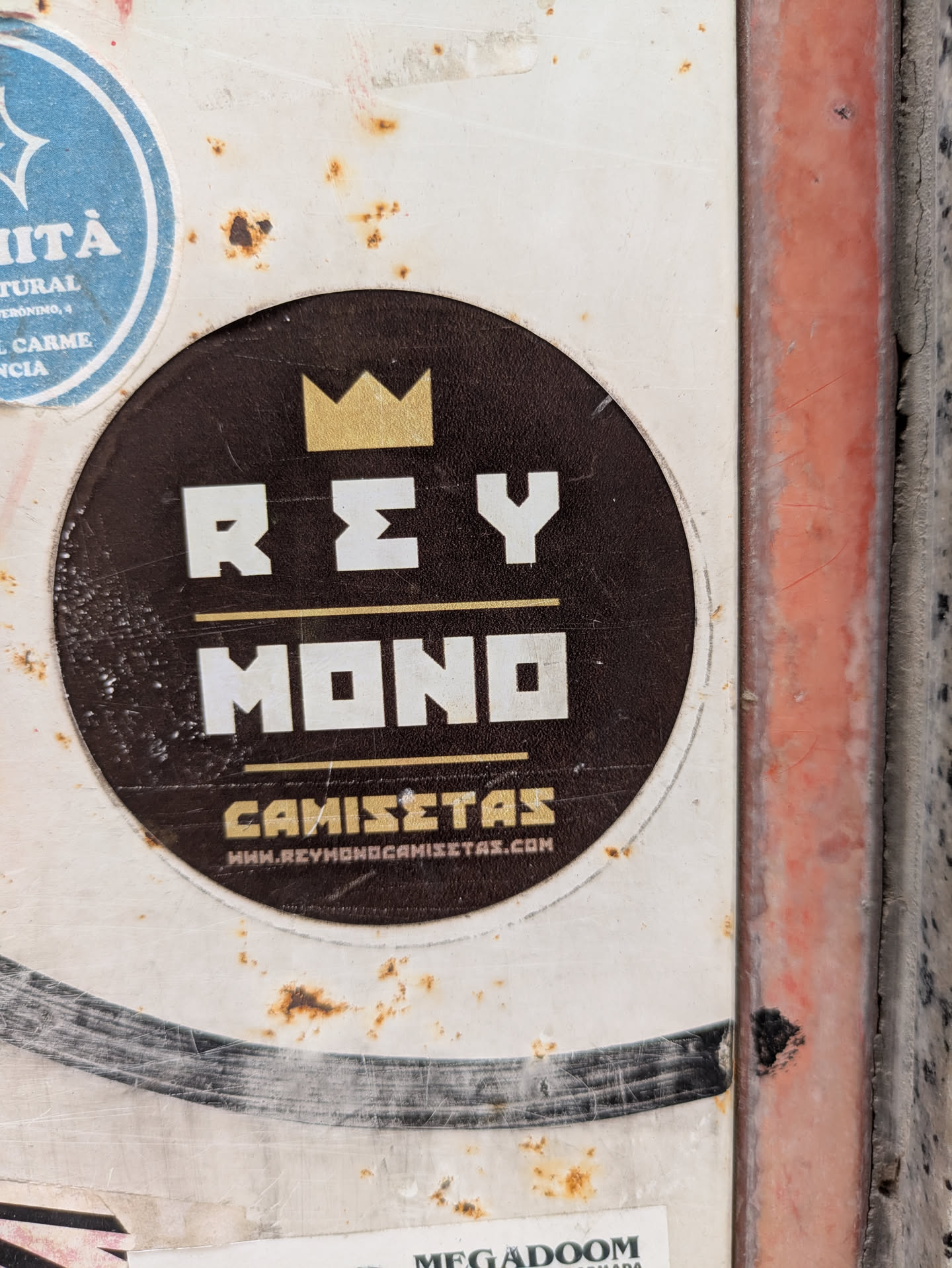 The sticker is round, with a dark brown background and a white border. At the top is a golden crown. Below the crown, in white block letters, is the text "REY MONO". Underneath that is the word "CAMISETAS" in gold letters. At the bottom of the sticker is the URL "WWW.REYMONOCAMISTAS.COM" in white letters.