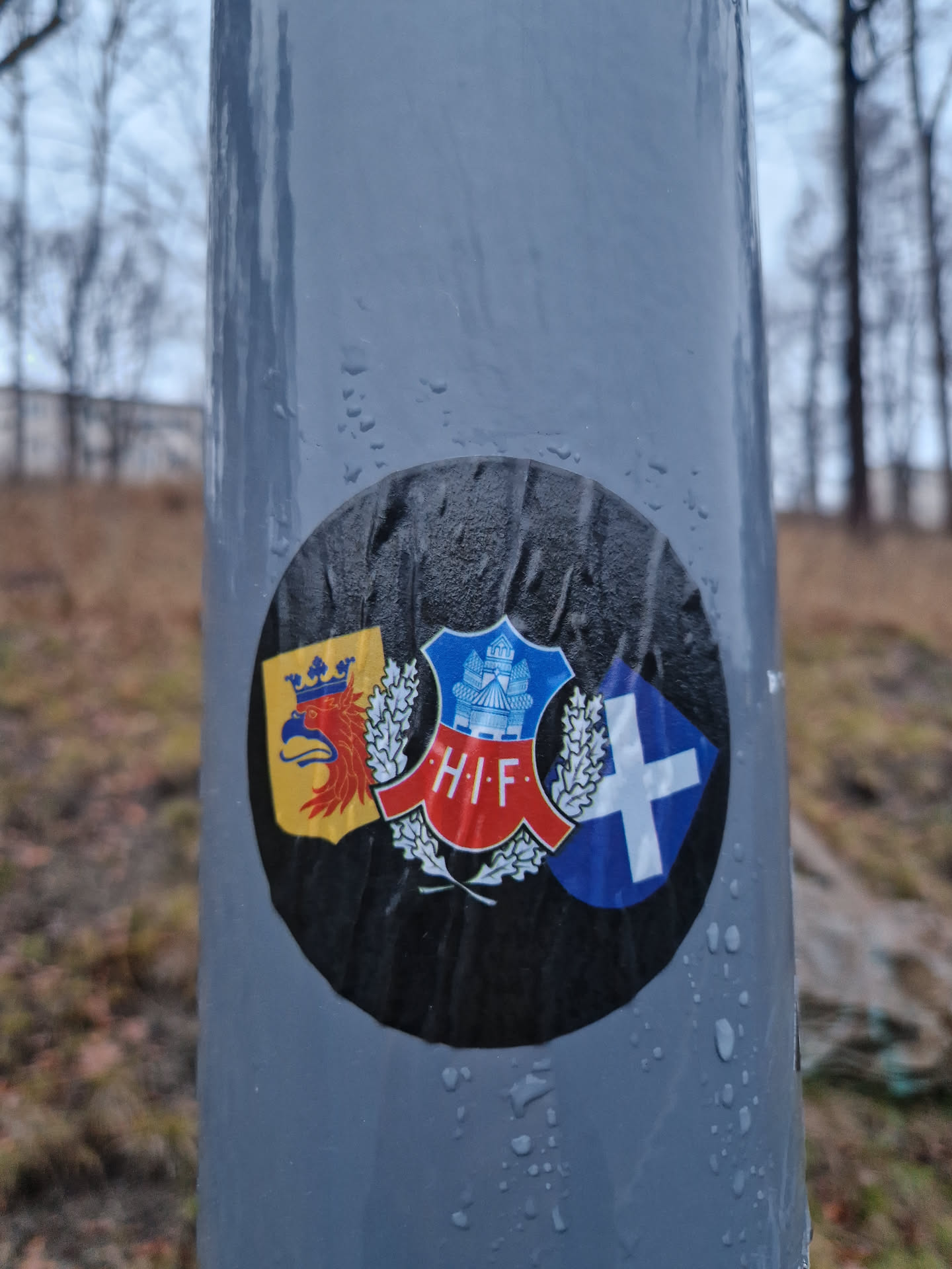 Round black sticker with three emblems. The central emblem is H.I.F. with wheat. On the left is a yellow emblem with an eagle and crown. On the right is a blue emblem with a white cross. The sticker is attached to a grey pole.