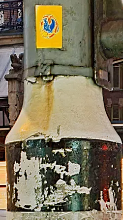 The image shows a weathered light post with peeling paint and a distinctive yellow sticker attached to its side. The sticker displays a blue and red abstract design that resembles a bird or flame. The post itself exhibits signs of age and wear, with various layers of paint flaking off, revealing the colors beneath. The surrounding environment includes architectural details of buildings in the background.