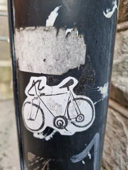 A black and white sticker featuring a drawing of a bicycle affixed to a dark-colored pole. The sticker shows some signs of wear and tear.