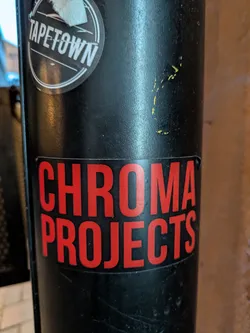 The sticker is rectangular with rounded corners and features large, sans-serif lettering in a vibrant red color against a black background. The text is divided into two lines. The top line reads 'CHROMA' and the bottom line reads 'PROJECTS'. The sticker appears to be adhered to a black cylindrical object, possibly a pole or a pipe. There is a slight peel or damage visible on the bottom right corner of the sticker.